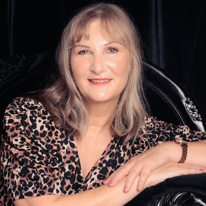 A portrait of a woman with shoulder-length blonde hair, smiling gently. She is wearing a leopard-print blouse and has her arms crossed in front of her, resting on a black, ornate chair. The background is a dark fabric, creating a soft contrast with her light skin and patterned blouse.